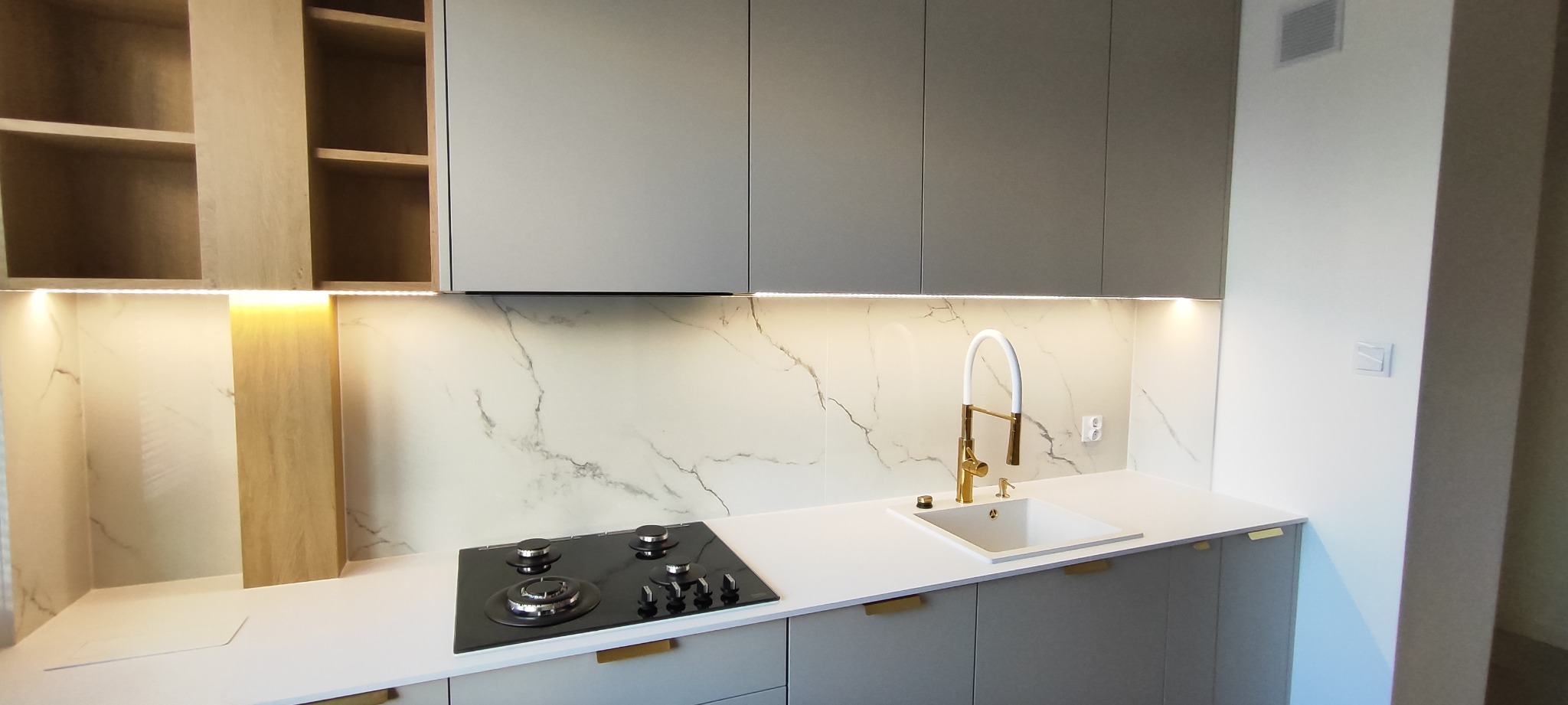Nowoczesna kuchnia z szarymi szafkami, marmurowym blatem i złotą armaturą. Oświetlenie LED podkreśla elegancki design. Widoczna płyta grzewcza i zlewozmywak.