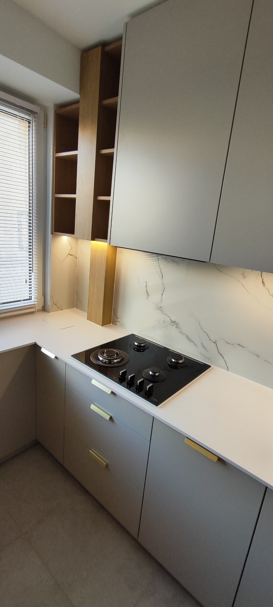 Nowoczesna kuchnia z szarymi szafkami, białym blatem i płytą indukcyjną. Oświetlenie LED podkreśla marmurowy wzór na ścianie. Widoczne otwarte półki i żaluzje w oknie.