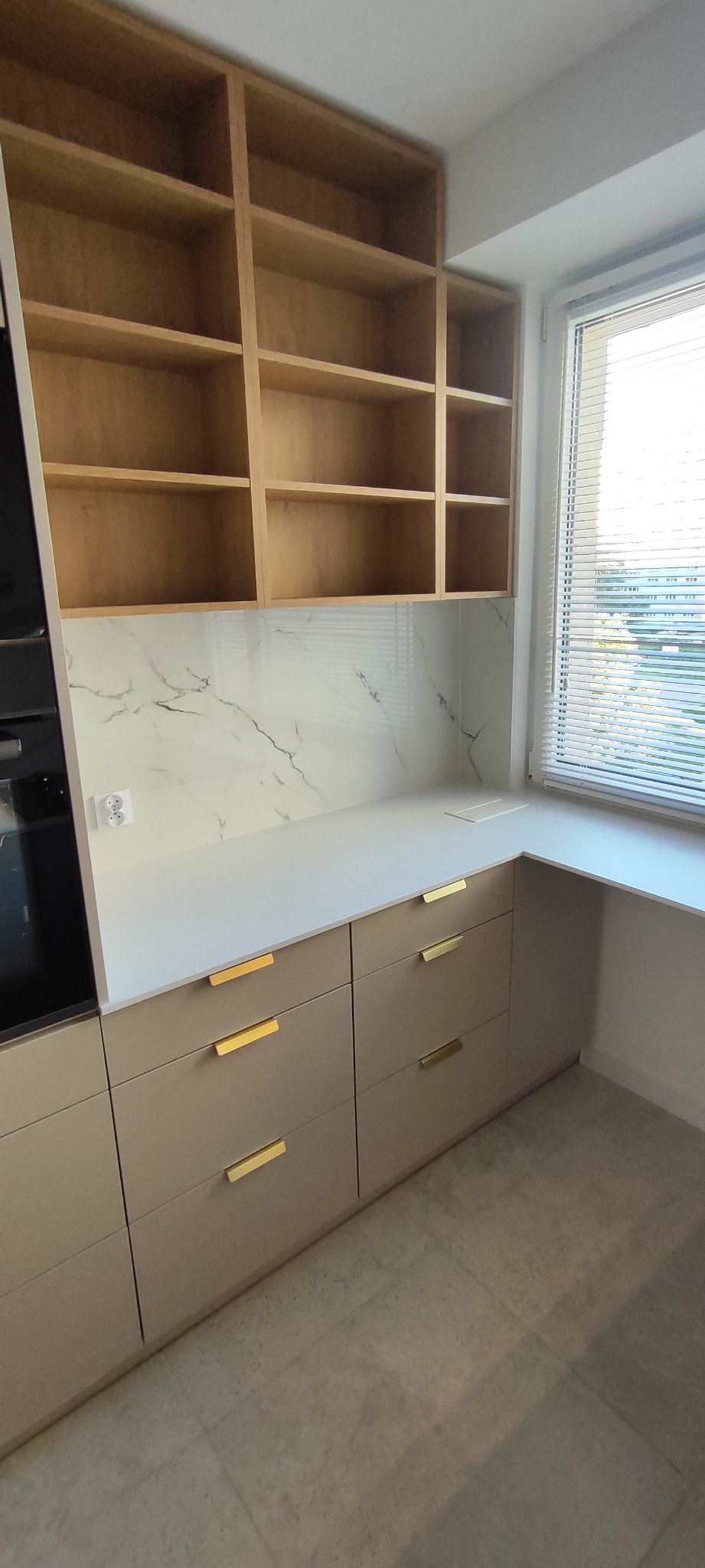 Beżowa zabudowa kuchenna z drewnianymi półkami, białym blatem i marmurowym panelem nad blatem. Złote uchwyty szuflad. Widok z góry na fragment kuchni.