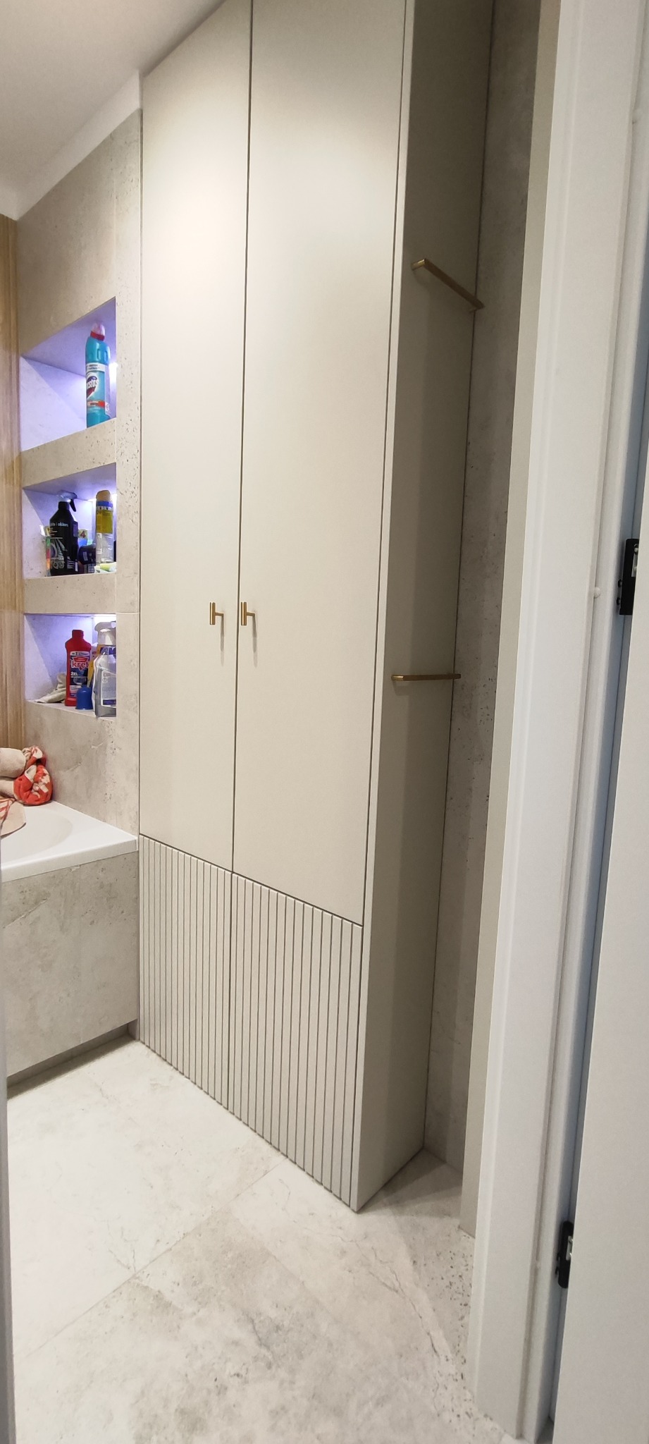 Biała szafa wnękowa z dekoracyjnym panelem w łazience, z półkami w ścianie i złotymi uchwytami, widok z perspektywy podłogi, jasne płytki podłogowe.