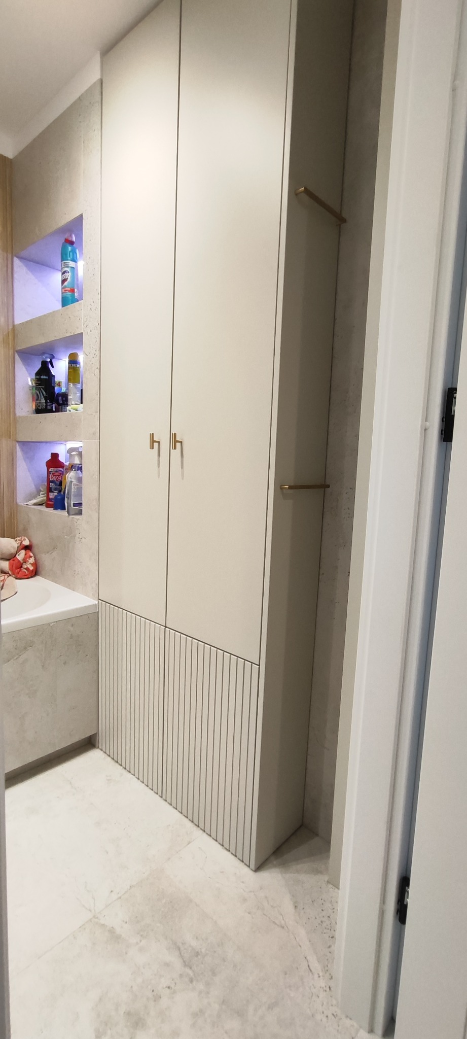 Biała szafa wnękowa z pionowymi żłobieniami w dolnej części, wkomponowana w aranżację łazienki z półkami i wanną obok.