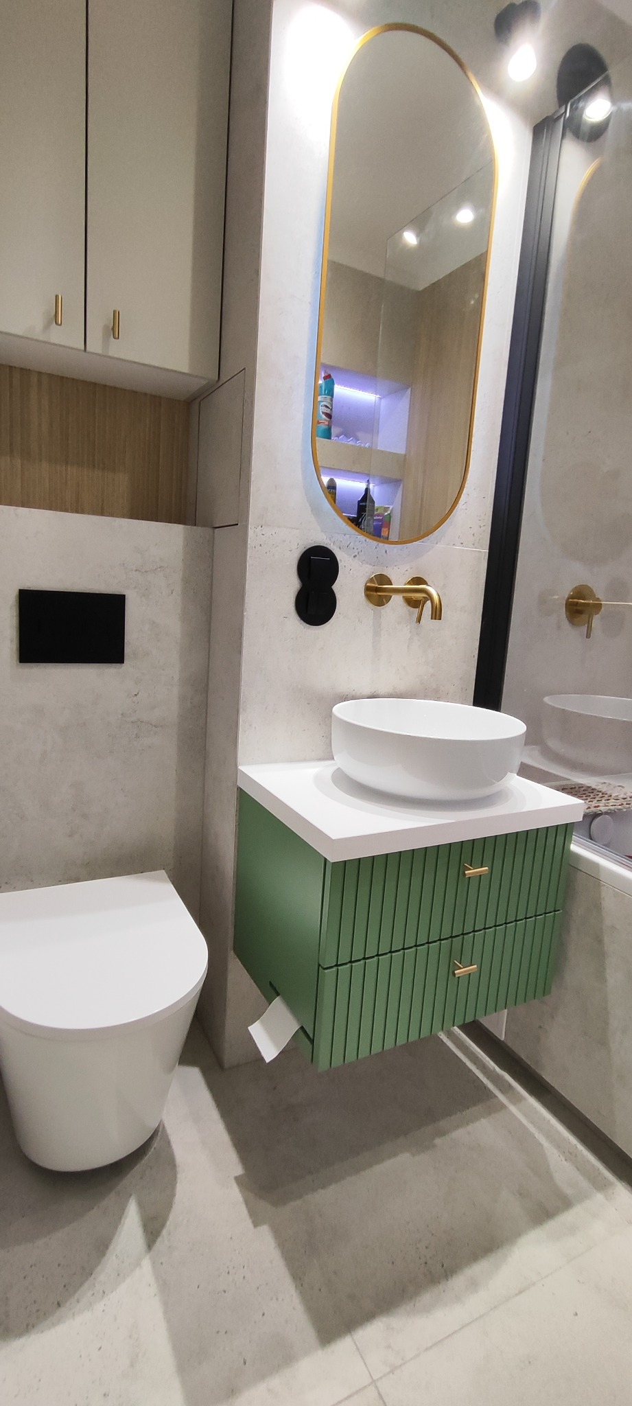 Nowoczesna łazienka z zieloną szafką pod umywalką, owalnym lustrem w złotej ramie i wiszącą toaletą. Minimalistyczny design, szare płytki, złote akcenty.