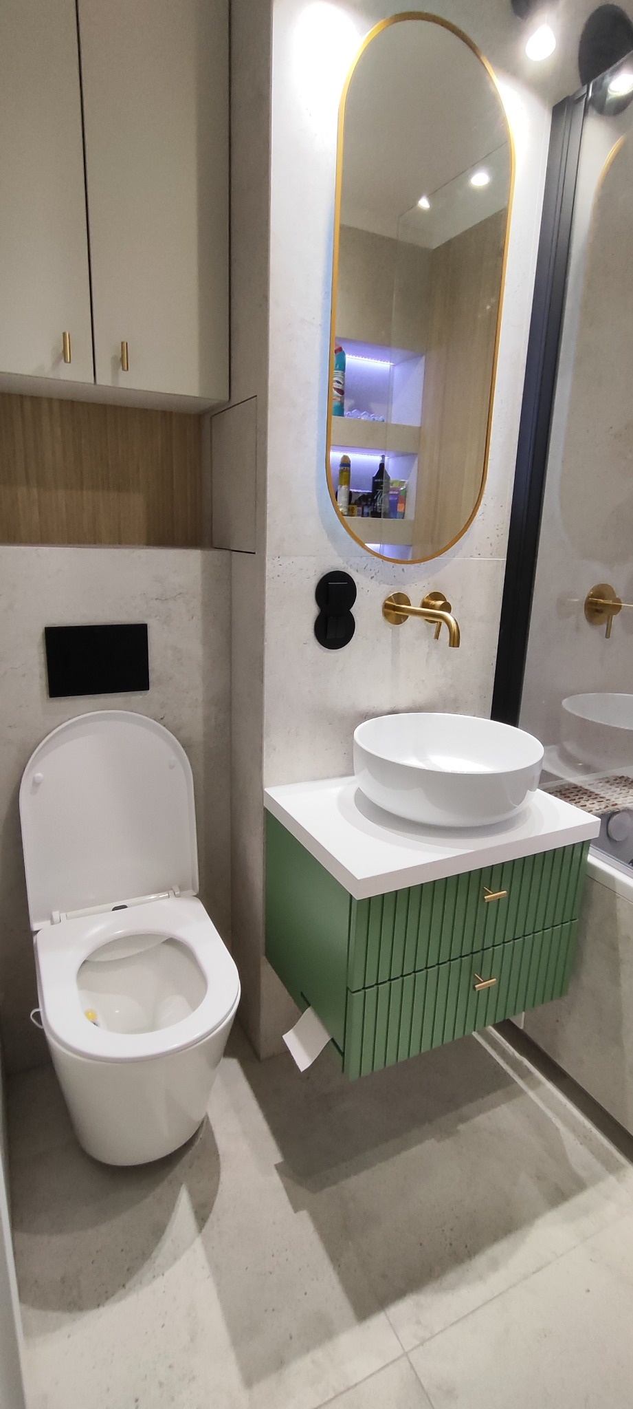 Nowoczesna łazienka z wiszącą toaletą, umywalką nablatową na zielonej szafce z frezowanymi frontami i owalnym lustrem w złotej ramie. Minimalistyczny design.