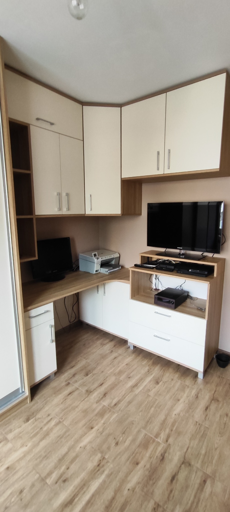 Funkcjonalna zabudowa meblowa w rogu pokoju: szafki wiszące, biurko z szafkami dolnymi i regał, tworzące spójną całość. Jasne fronty i drewnopodobny blat.