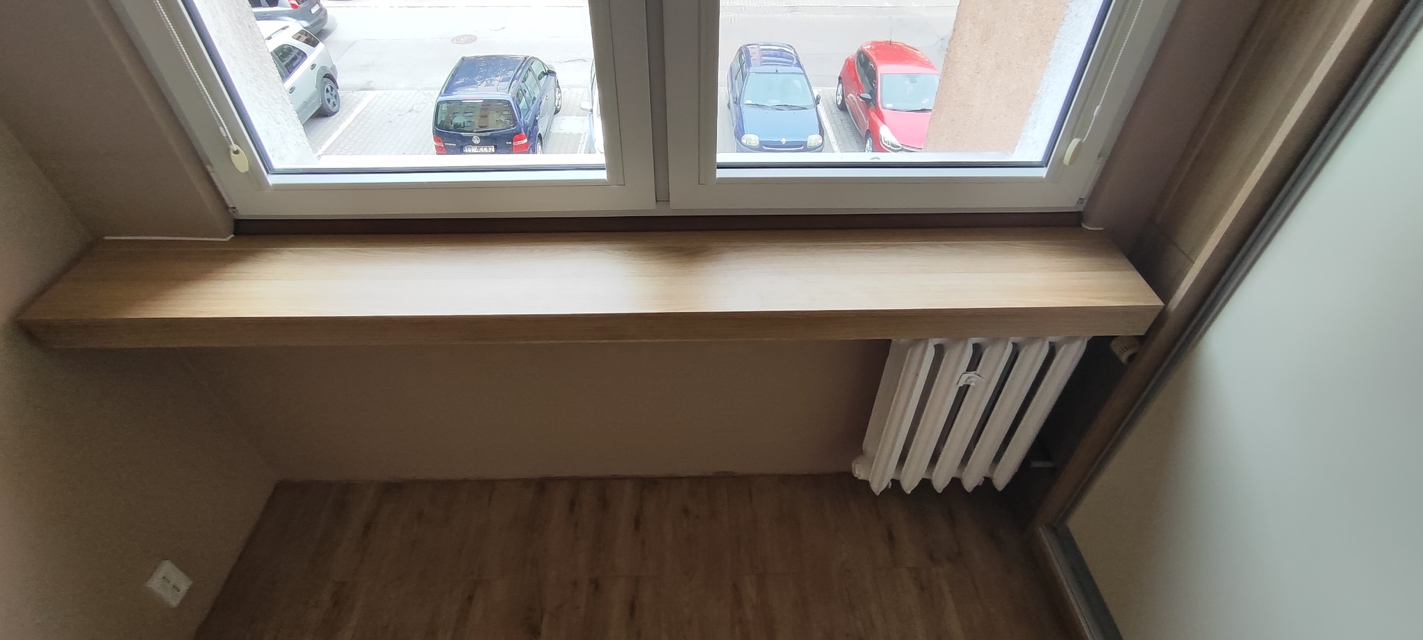Drewniany parapet z widokiem na parking w Łodzi. Jasne drewno, białe okno, grzejnik pod spodem. Ujęcie z góry, widoczne panele podłogowe.