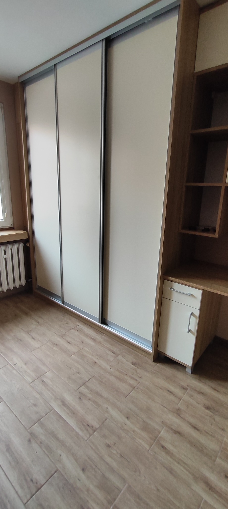Jasna szafa wnękowa z przesuwnymi drzwiami i biurkiem z półkami, dopasowana kolorystycznie do podłogi z paneli. Widok na całą konstrukcję w pokoju.