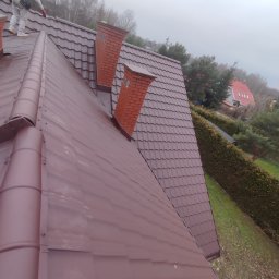 LEONID VASHCHENKO - Ciemnobrązowy dach z blachodachówki trapezowej, dwa kominy z cegły, pracownik w kombinezonie zabezpieczającym. Widok z góry na dom w tle, pochmurny dzień.