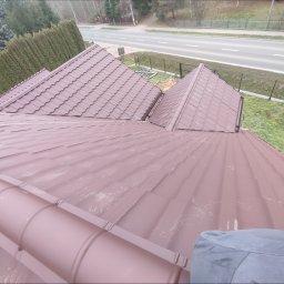 LEONID VASHCHENKO - Widok z góry na nowy, brązowy dach z blachodachówki modułowej, z widocznymi elementami mocującymi i fragmentem osoby w prawym dolnym rogu. W tle droga i zieleń.