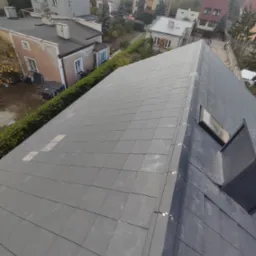 Grafitowy dach z dachówki karpiówki, widok z góry na tle zabudowy miejskiej i zieleni. Nowe pokrycie z oknem dachowym, zabezpieczenia na krawędziach.