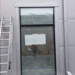 LEONID VASHCHENKO - Szara fasada budynku z oknem zabezpieczonym folią i aluminiową drabiną. Widoczna rynna spustowa. Detal elewacji przemysłowej w trakcie prac.