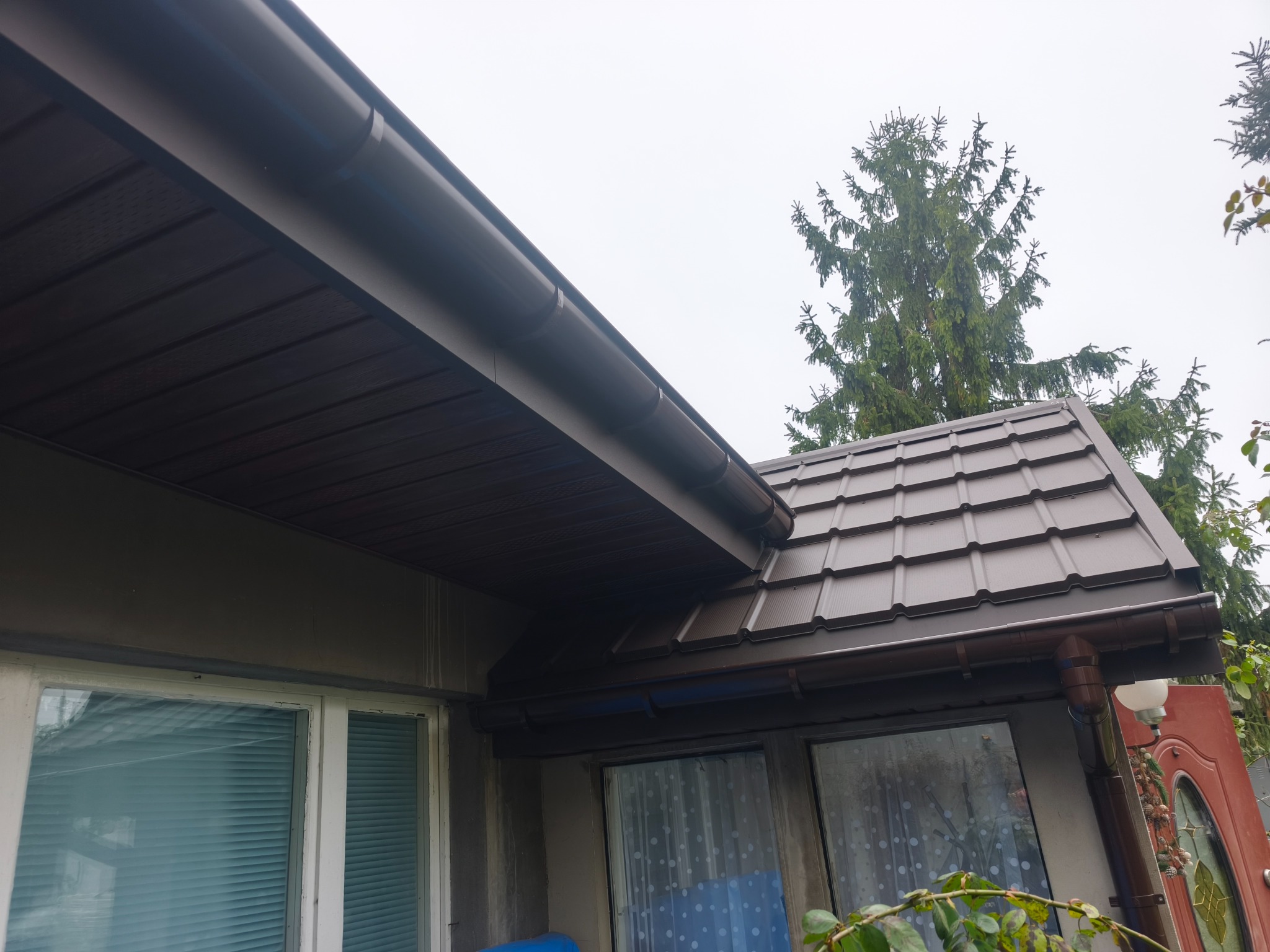 Brązowy dach z blachy trapezowej i rynny na tle szarej elewacji domu oraz zielonego drzewa iglastego. Widok z dołu, podkreślający detale wykończenia.