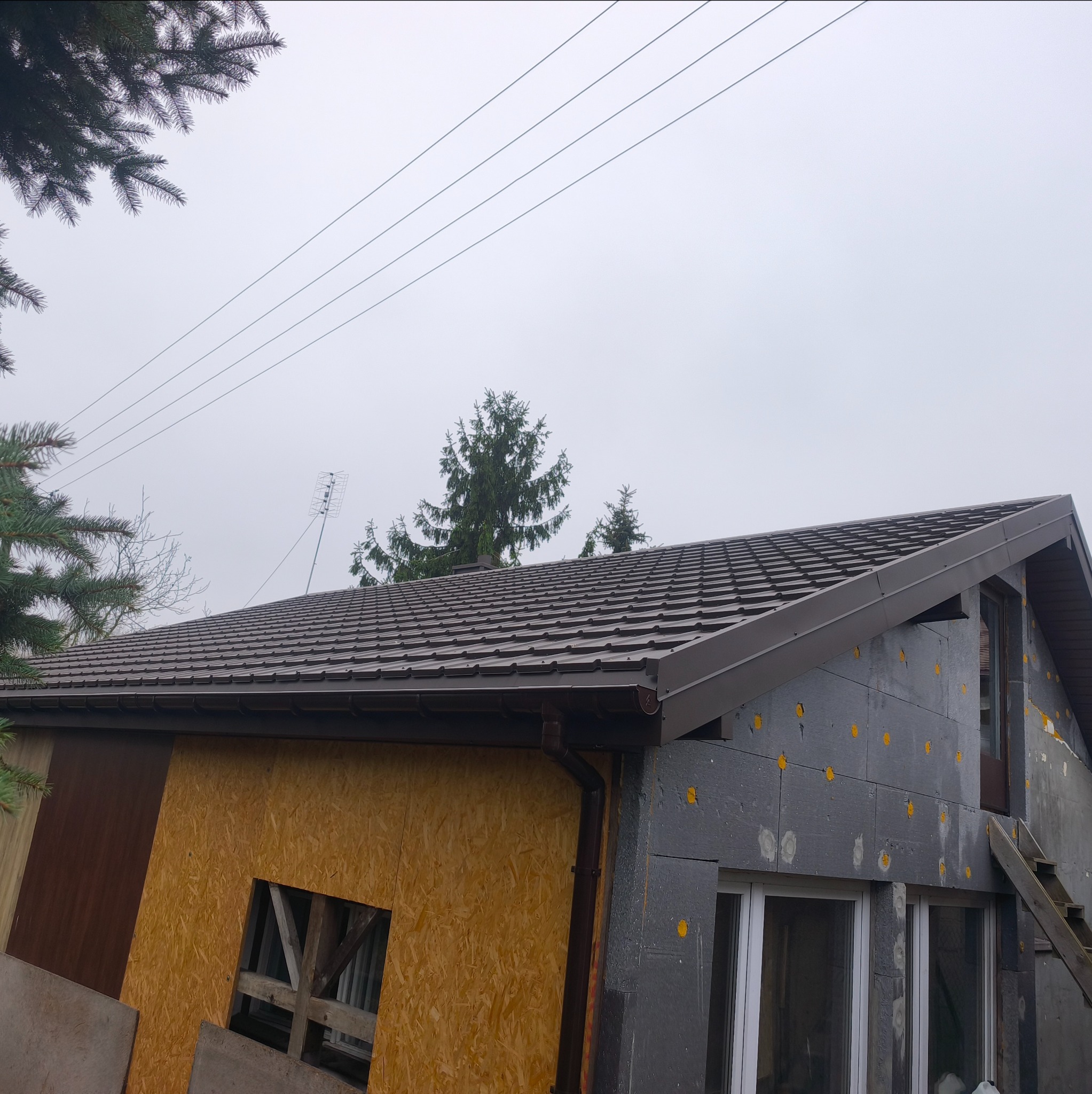 Dom w trakcie prac dekarskich: brązowy dach z blachy trapezowej, ocieplenie ścian styropianem grafitowym, widoczne rynny i fragmenty elewacji z desek.