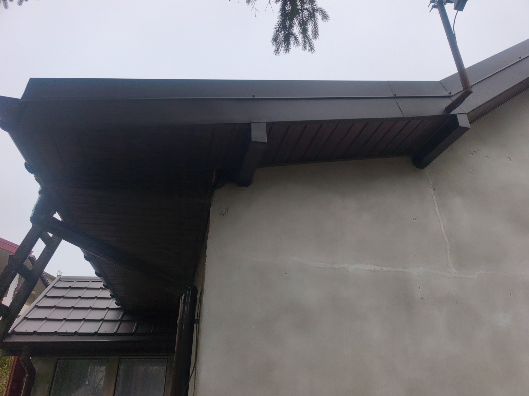 Fragment brązowego dachu z rynną i podbitką, widoczny z dołu pod kątem. Na ścianie budynku widoczne ślady po naprawach. W tle fragment dachu sąsiedniego budynku.