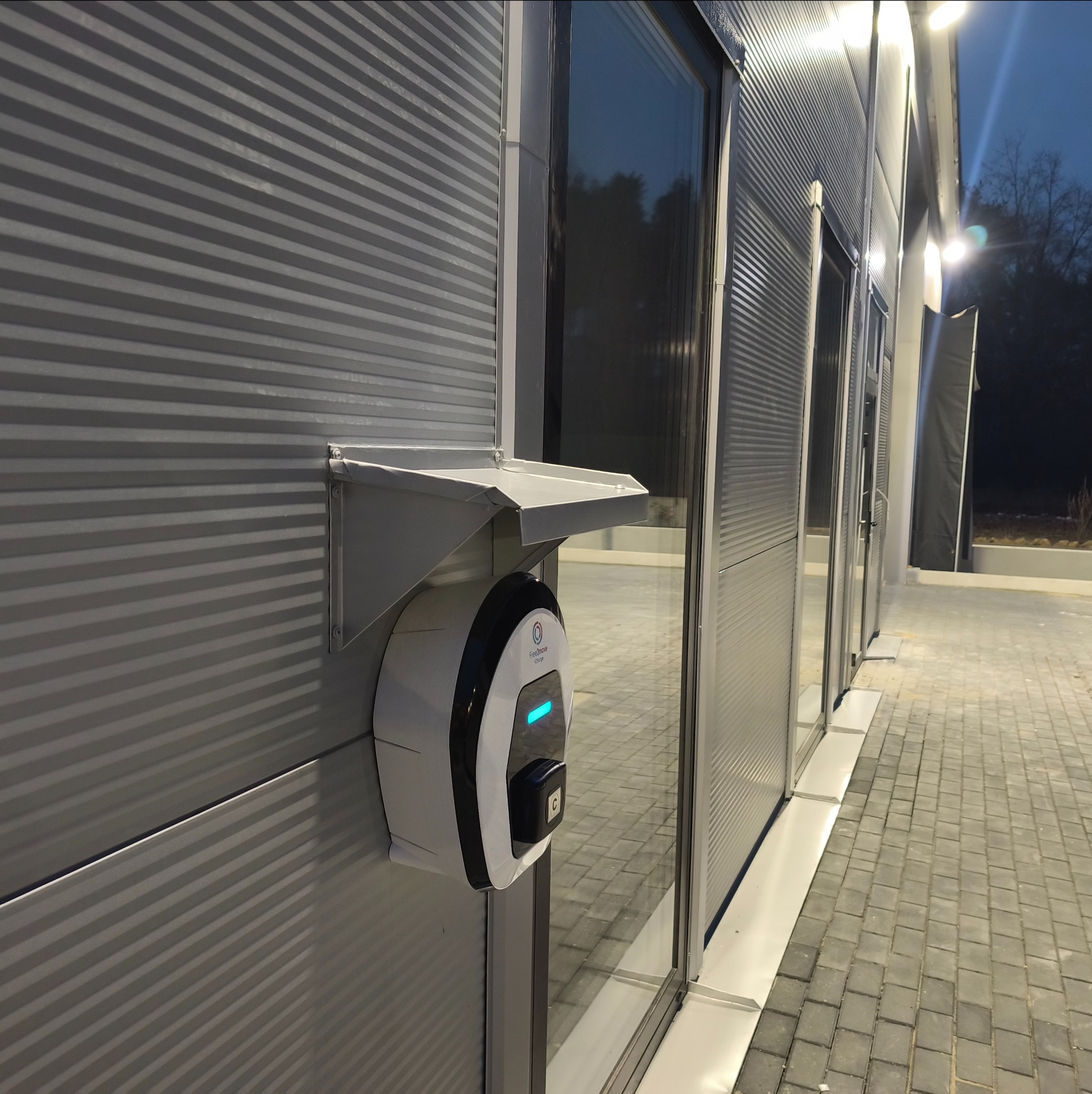 Nowoczesna stacja ładowania pojazdów elektrycznych zamontowana na elewacji budynku z blachy trapezowej, oświetlona światłem zewnętrznym.
