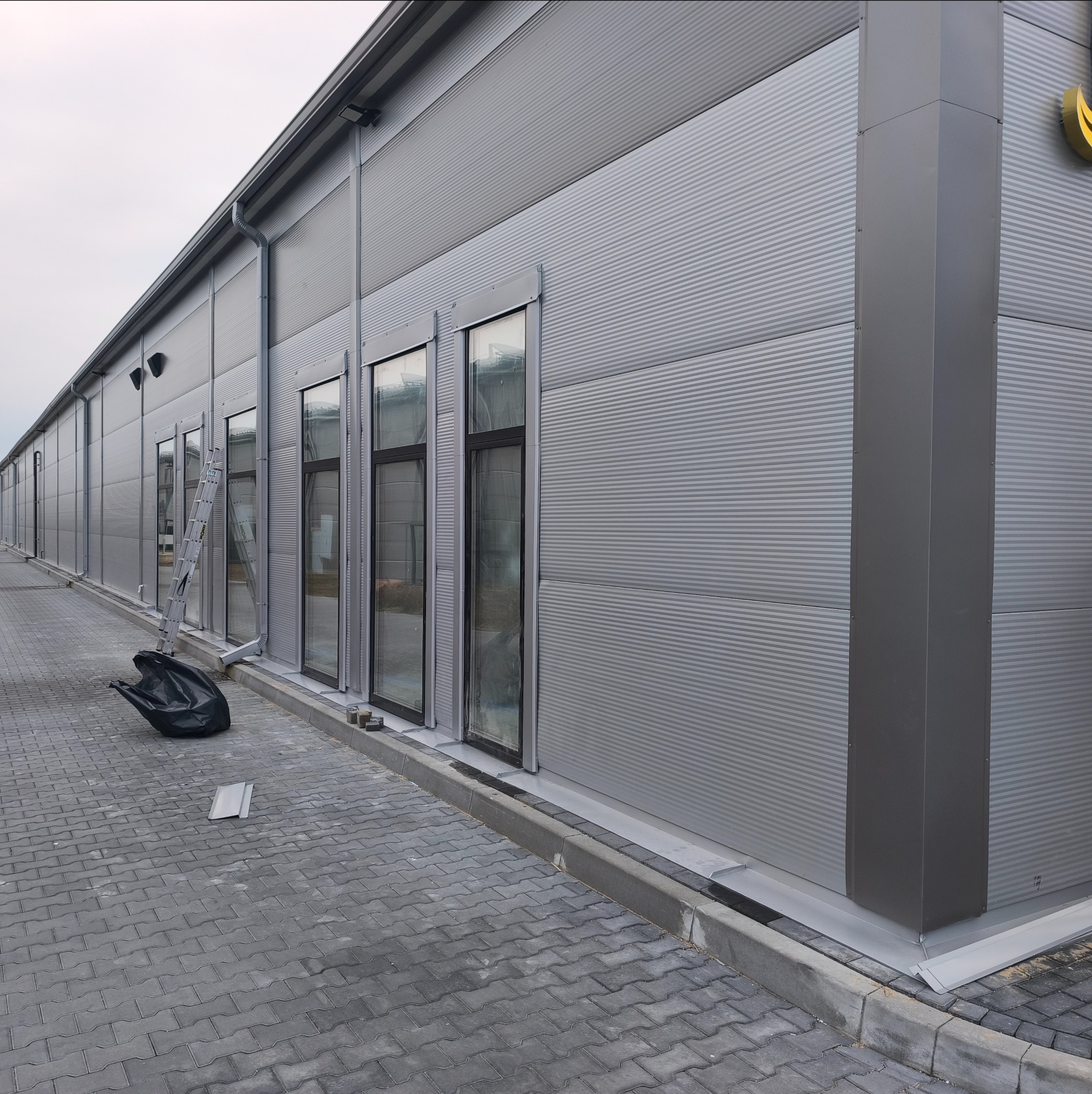 Szary budynek przemysłowy z blachy trapezowej, widoczne okna, rynny, drabina oparta o ścianę i czarny worek na chodniku. Ujęcie z dołu, pochmurne niebo.