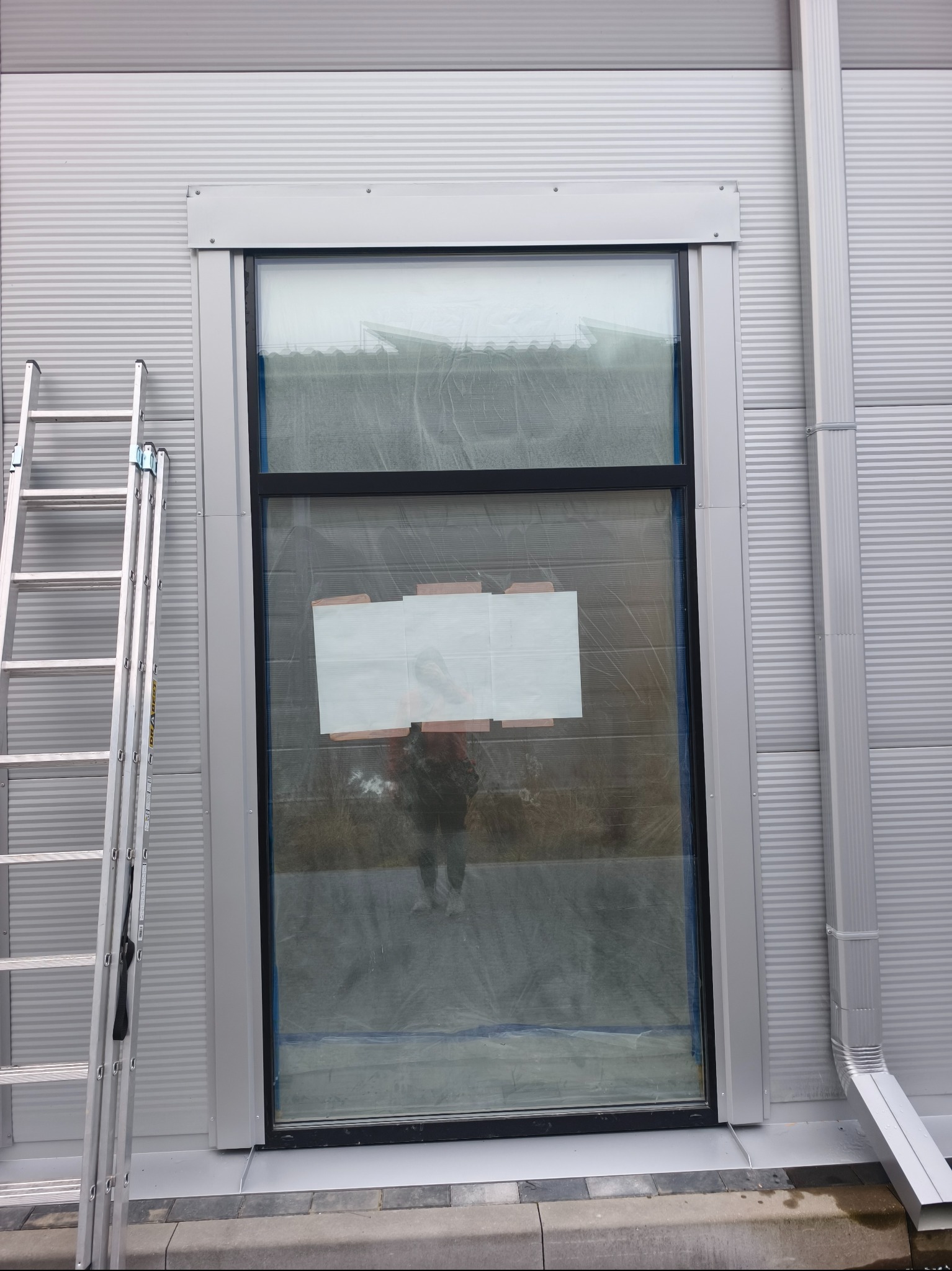 Szara fasada budynku z oknem zabezpieczonym folią i aluminiową drabiną. Widoczna rynna spustowa. Detal elewacji przemysłowej w trakcie prac.