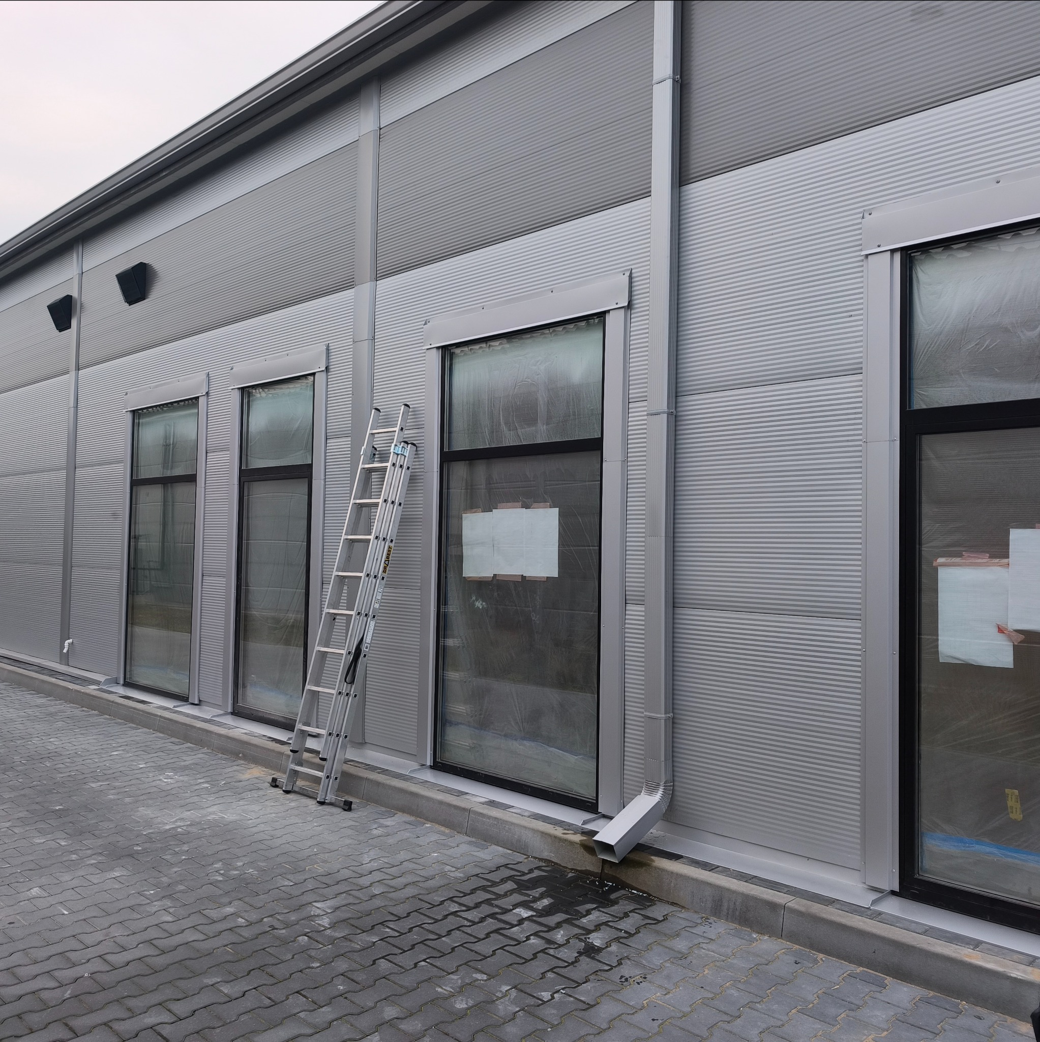 Szara, metalowa elewacja budynku z rynnami i oknami zabezpieczonymi folią. Aluminiowa drabina oparta o ścianę. Widok z zewnątrz na kostkę brukową.