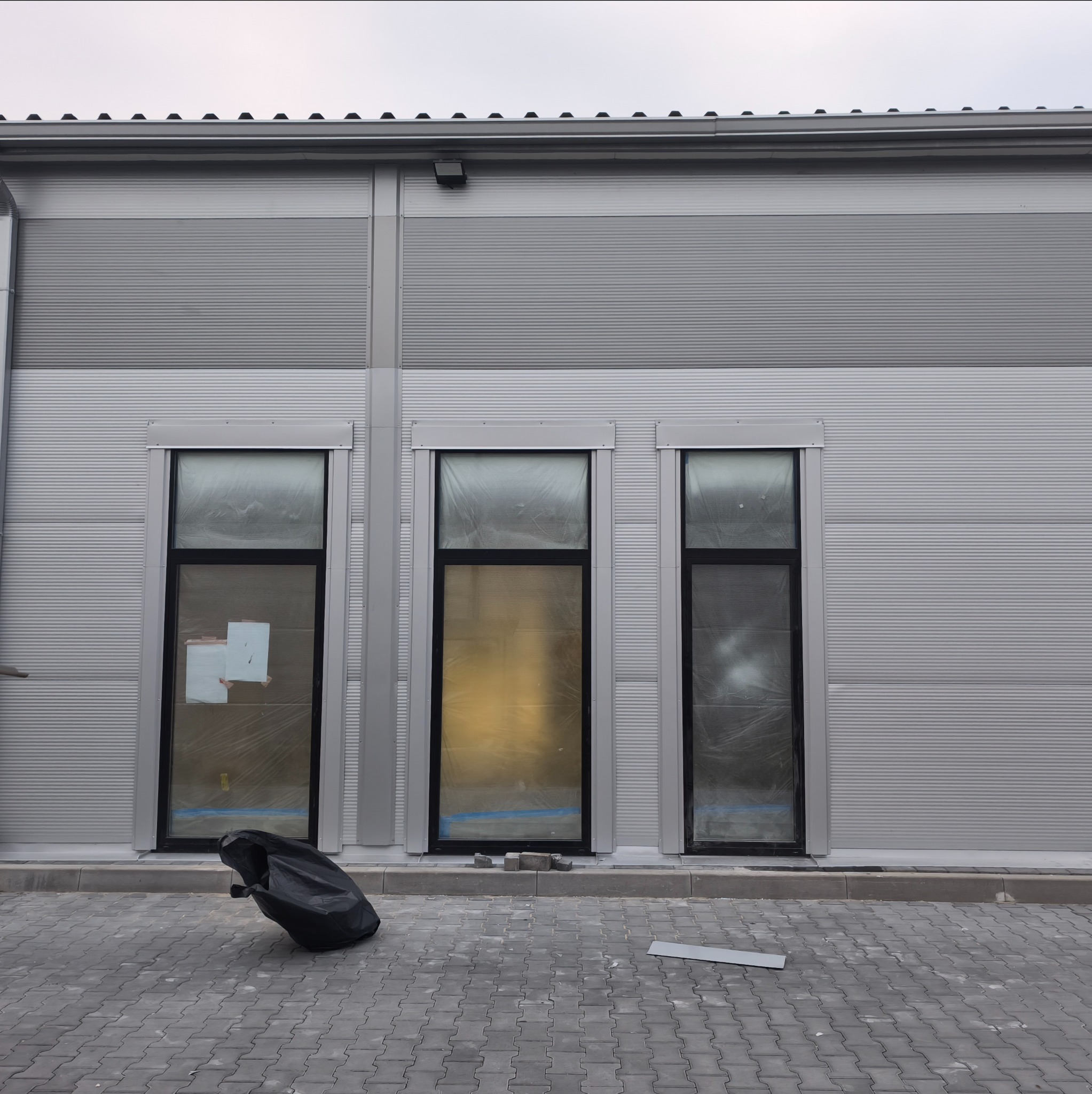 Szara, blaszana elewacja budynku z trzema oknami zabezpieczonymi folią. Na chodniku leży czarny worek i fragment blachy. Widoczne elementy dachu.