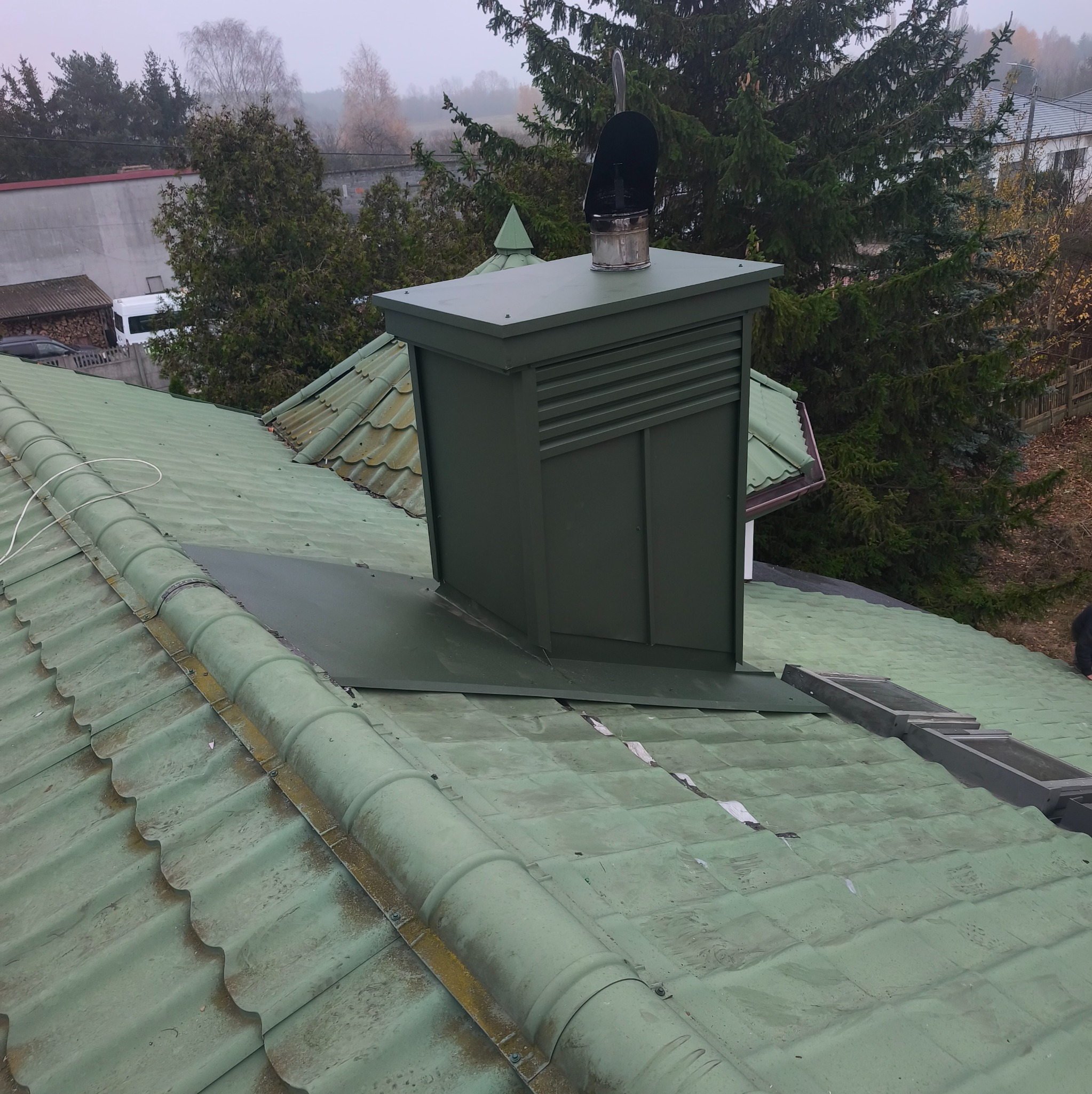Zielony komin wentylacyjny na dachu z zielonej dachówki ceramicznej. Widoczne okna dachowe i drzewa w tle. Dach pokryty mchem, co sugeruje potrzebę czyszczenia.