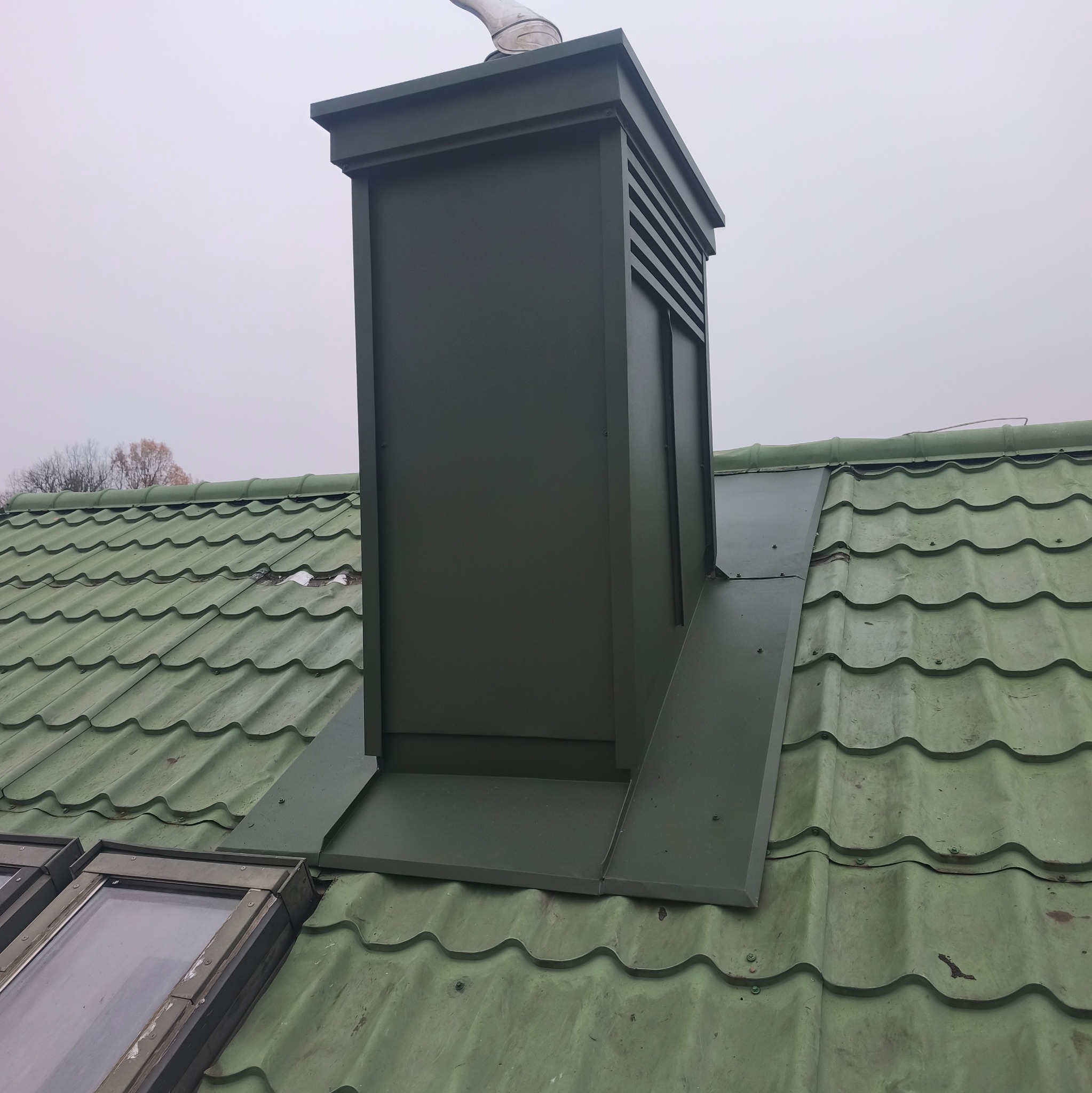 Zielony komin wentylacyjny z obróbką blacharską na dachu z zielonej blachodachówki. Widoczne okna dachowe i fragment drzewa w tle.