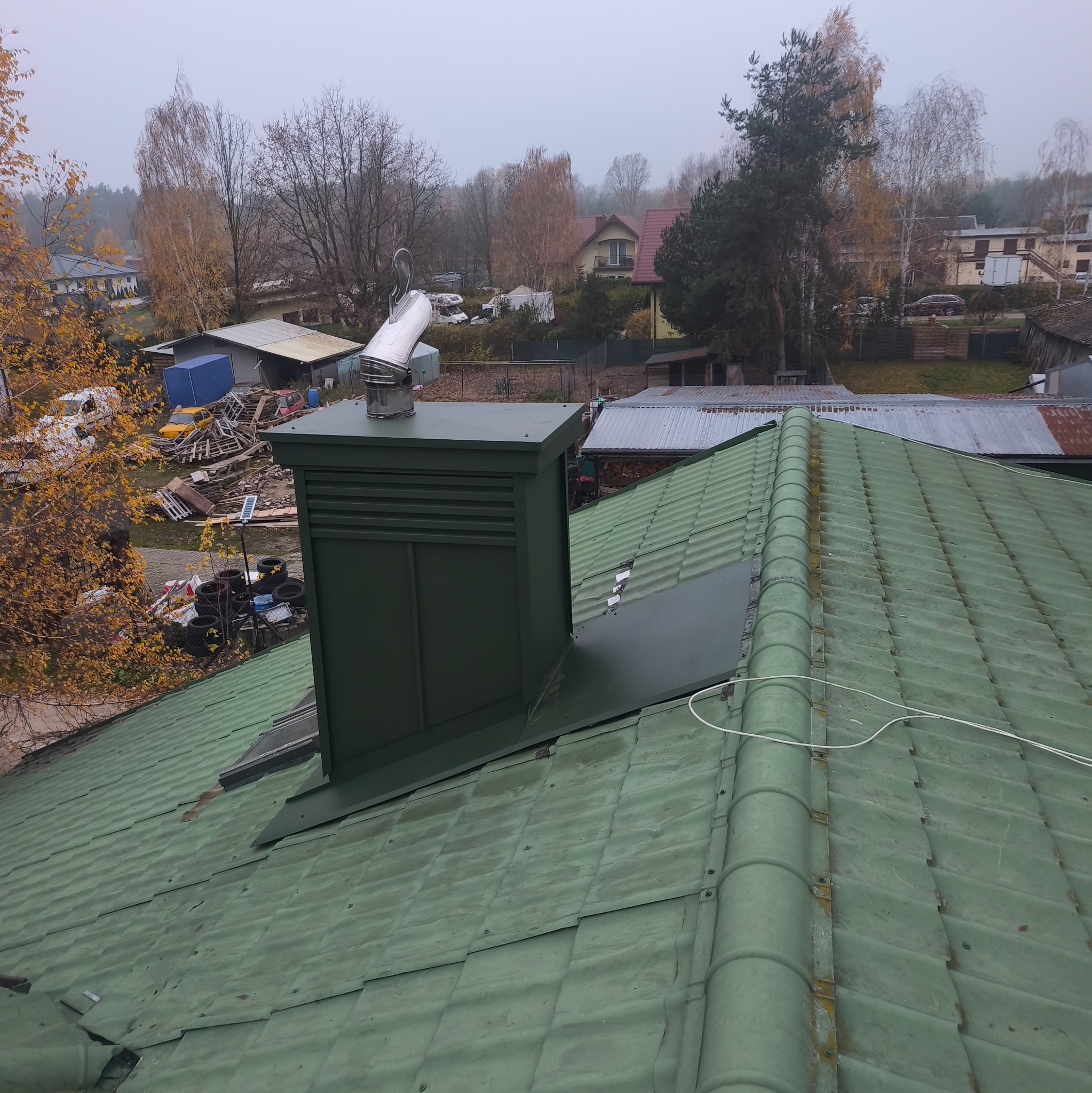 Zielony dach z kominem wentylacyjnym na tle jesiennego krajobrazu. Widoczne uszkodzenia i zabrudzenia na pokryciu dachowym. Komin w kolorze dachu.