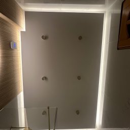 MF-PERFECTO Mateusz Filipek - Nowoczesna łazienka z kabiną prysznicową, widok od dołu na podwieszany sufit z sześcioma reflektorami i liniowym oświetleniem LED. Ściana z drewnianymi lamelami, złota deszczownica.