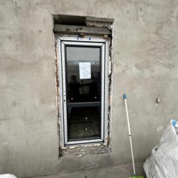 Burzkurz - Montaż okna w surowej ścianie z widocznymi szczelinami i pianką montażową. Obok ściany stoi miotła i worki. W szybie okna odbija się osoba.