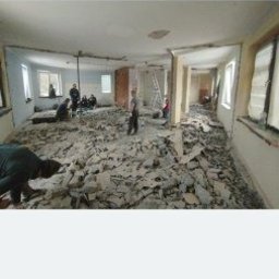 Burzkurz - Wnętrze w trakcie prac: gruz na podłodze, ściany częściowo usunięte, ekipa w tle. Widoczne okna i resztki instalacji. Ujęcie z perspektywy osoby stojącej.