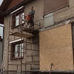 Burzkurz - Pracownik na rusztowaniu, w kasku, używa młota pneumatycznego do pracy na elewacji budynku. Część okien zabita płytami. Widoczna drabina i ogrodzenie.