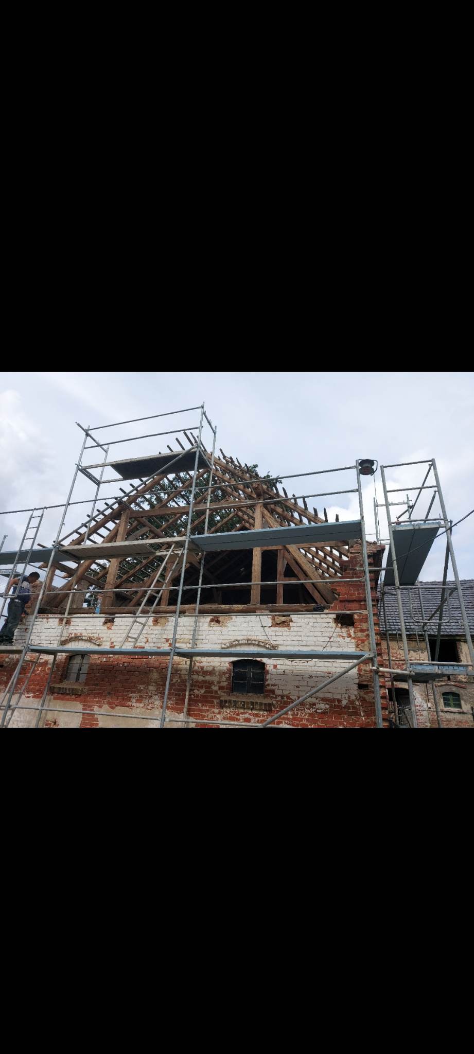 Ceglany budynek w trakcie rozbiórki dachu, widoczne rusztowania i elementy konstrukcyjne. Częściowo otynkowana ściana z oknami. Prace na wysokościach.