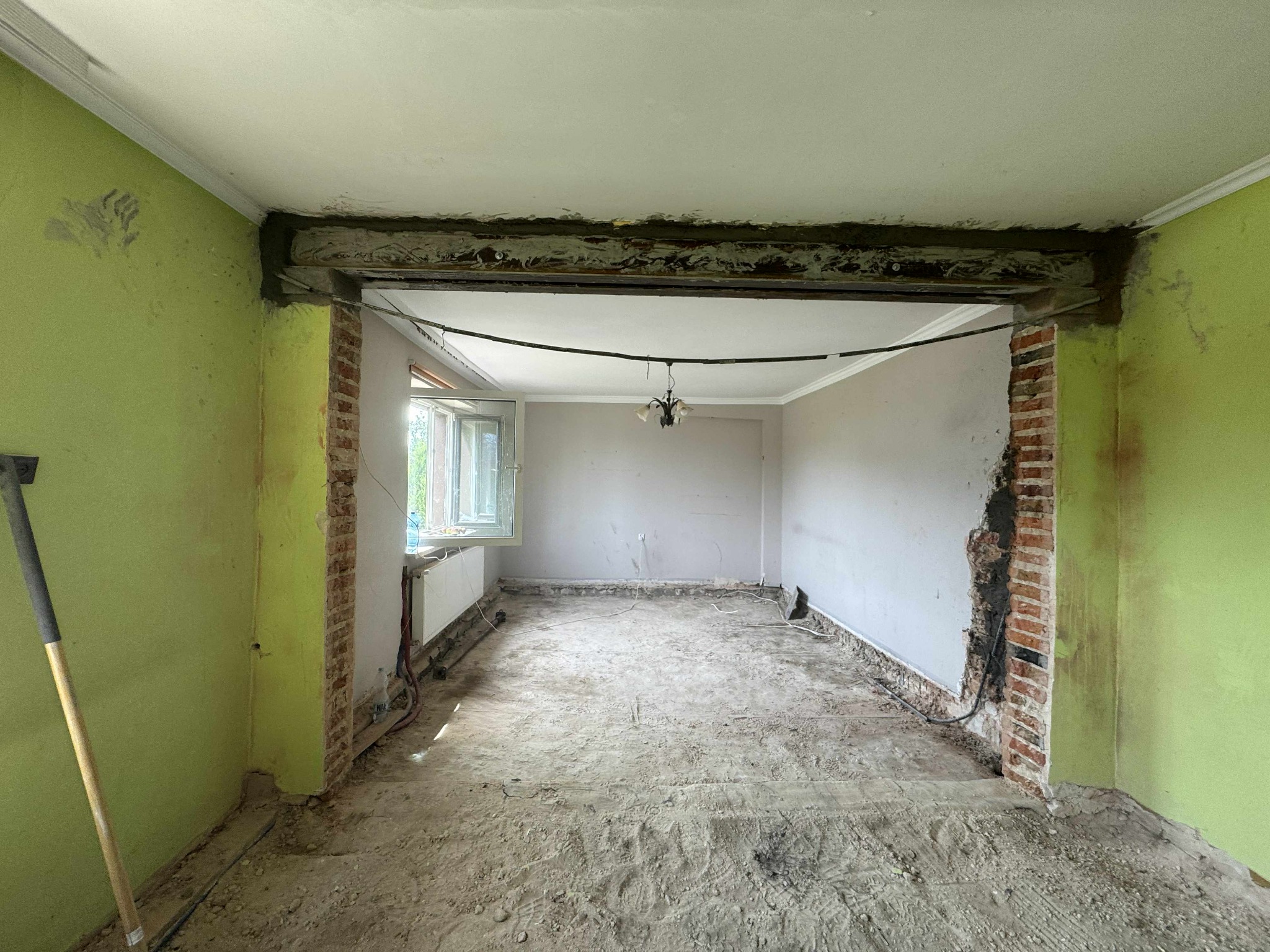 Wnętrze po wyburzeniu ściany działowej, widoczne odsłonięte cegły, resztki gruzu na podłodze i ślady po instalacjach. Ściany w kolorze zielonym i szarym.