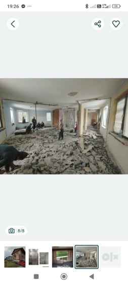 Wnętrze w trakcie prac: gruz na podłodze, ściany częściowo usunięte, ekipa w tle. Widoczne okna i resztki instalacji. Ujęcie z perspektywy osoby stojącej.