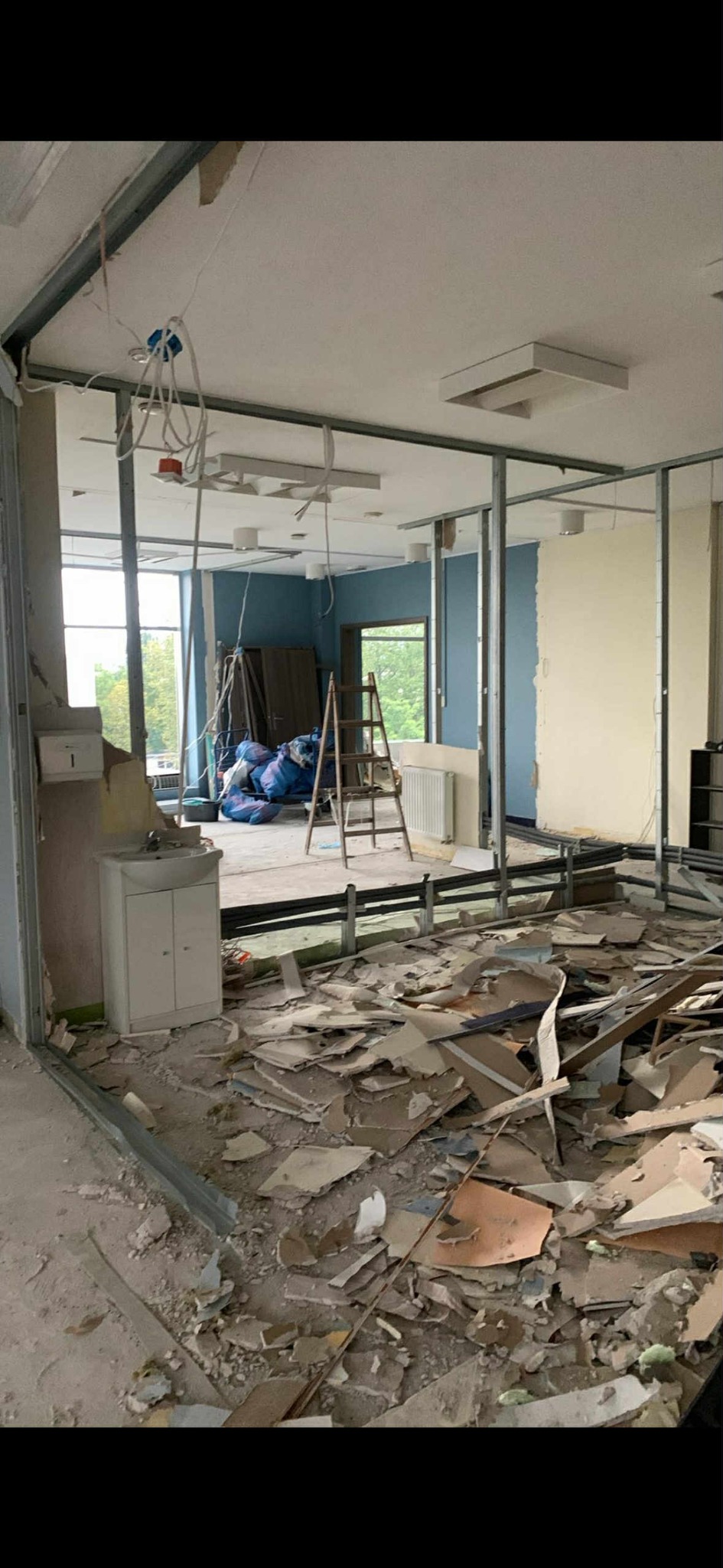 Wnętrze w trakcie wyburzania ścian działowych. Widoczne gruz, odsłonięte instalacje, drabina i okno z widokiem na zieleń. Metalowe profile konstrukcyjne.