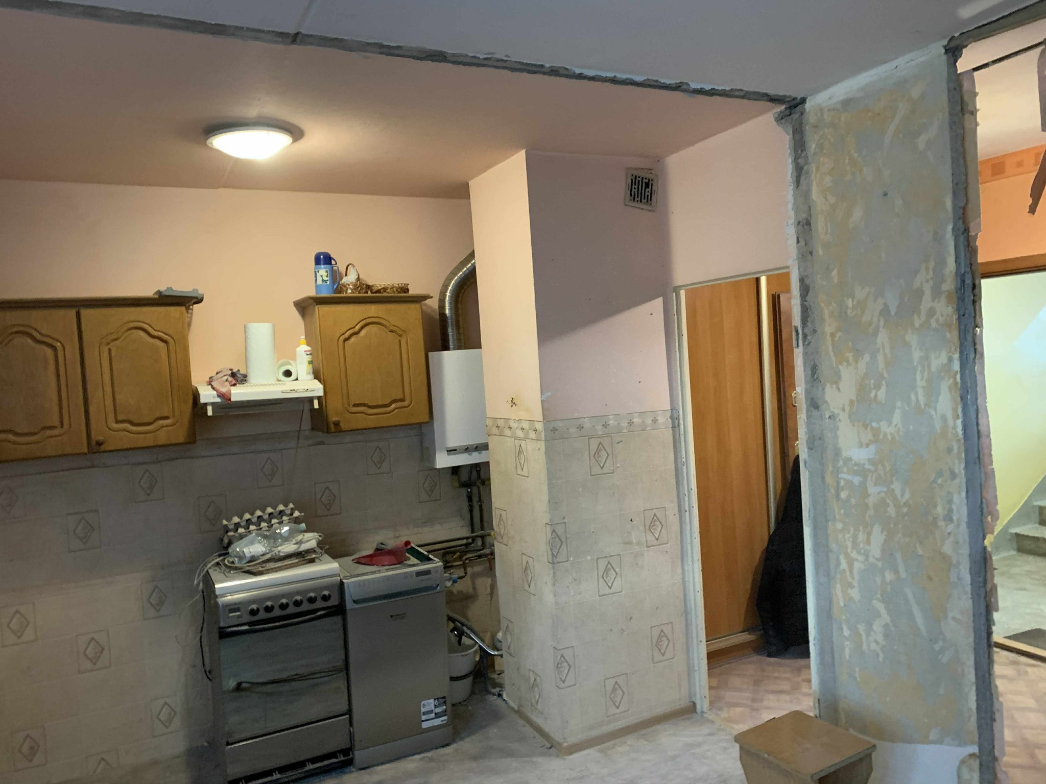 Kuchnia w trakcie remontu: częściowo usunięta ściana działowa, odsłonięte instalacje, widoczne meble kuchenne i sprzęty AGD. Ślady po wyburzeniach na ścianach i suficie.