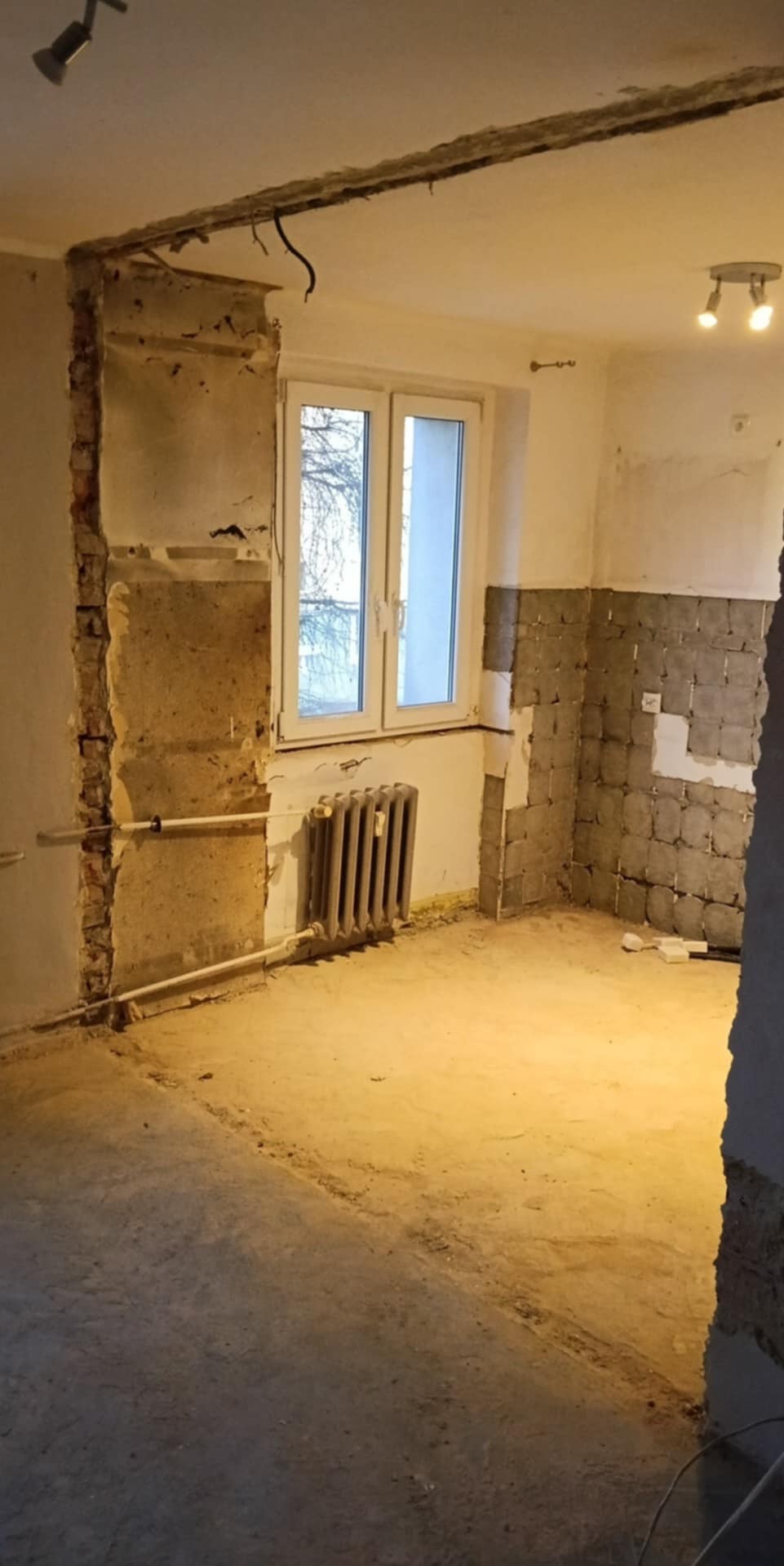 Wnętrze w trakcie demontażu: odsłonięte cegły, fragmenty płytek, grzejnik pod oknem. Widoczne ślady po instalacjach. Surowy stan, przygotowanie do remontu.