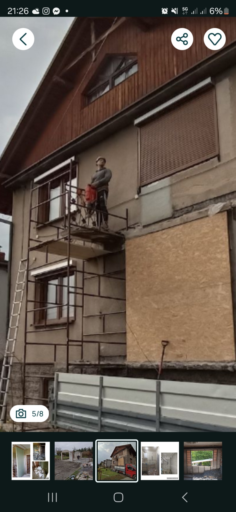 Pracownik na rusztowaniu, w kasku, używa młota pneumatycznego do pracy na elewacji budynku. Część okien zabita płytami. Widoczna drabina i ogrodzenie.
