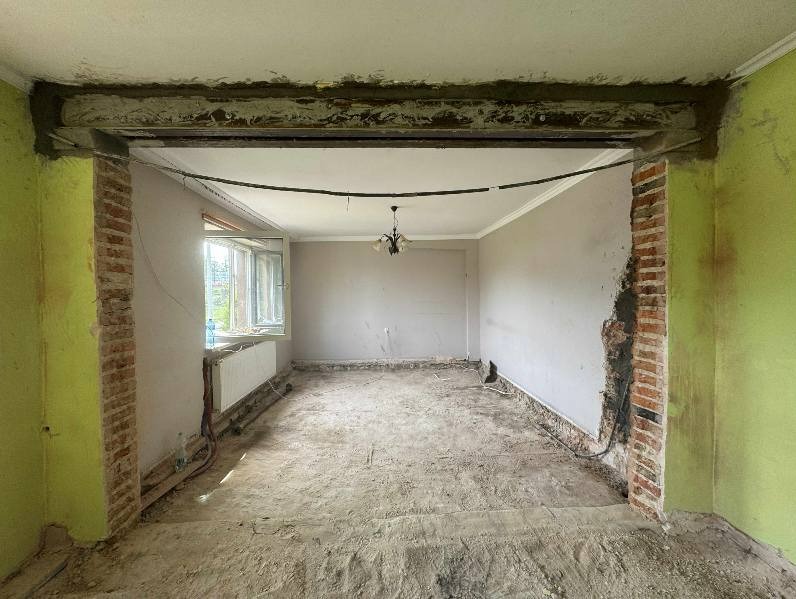 Wnętrze po wyburzeniu ściany działowej, widoczne odsłonięte cegły i pozostałości po konstrukcji nośnej. Ściany w kolorze zielonym i szarym, okno z grzejnikiem.