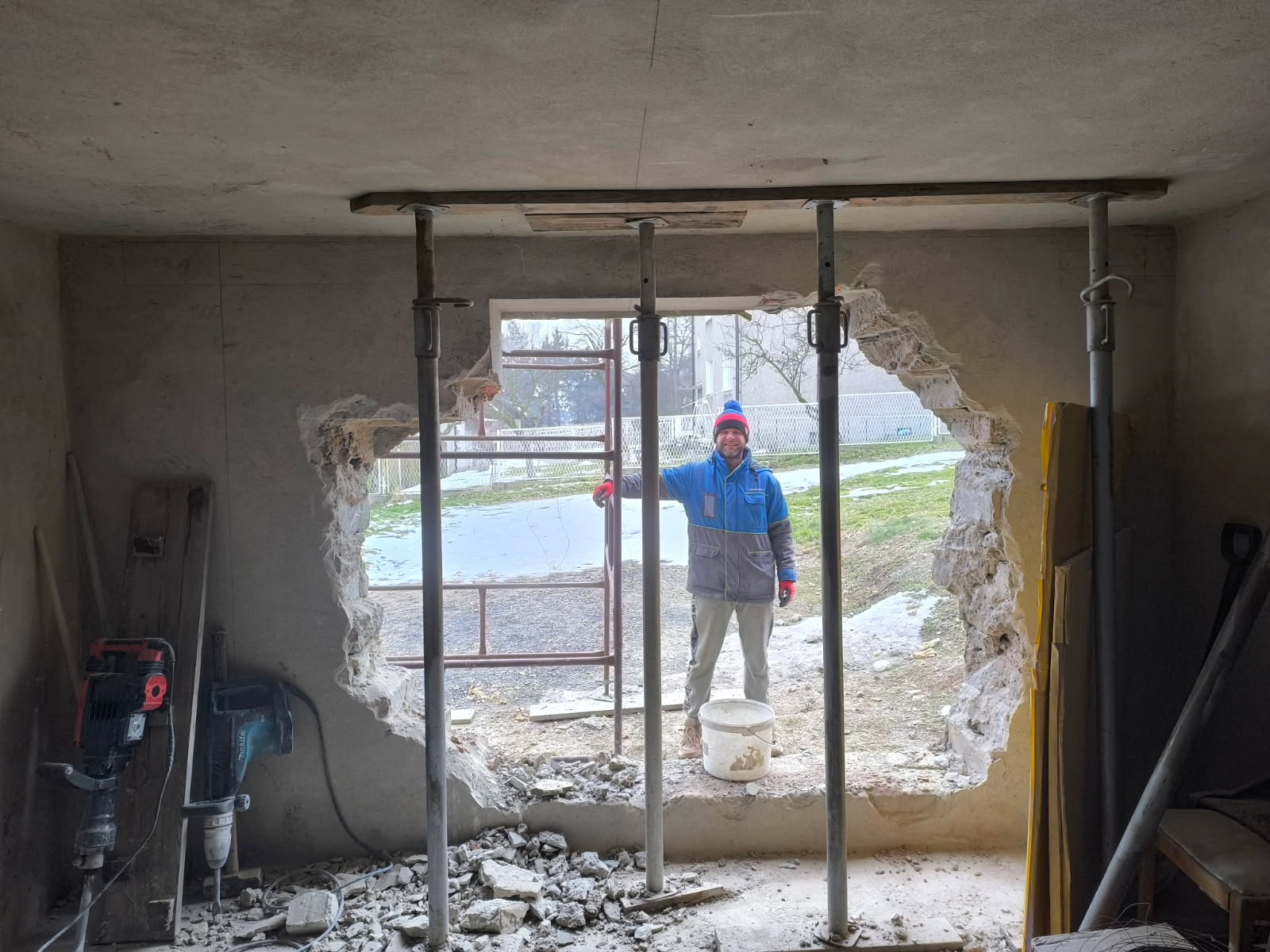 Otwór w ścianie podparty stemplami budowlanymi, widoczny pracownik w niebieskiej kurtce i narzędzia do wyburzania. Prace wewnątrz budynku, gruz na podłodze.