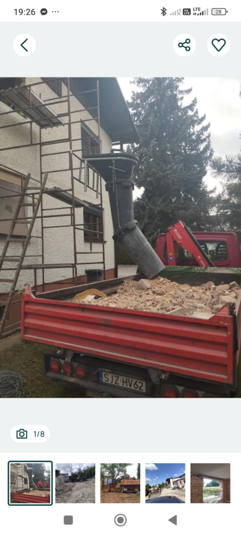 Czerwona ciężarówka wypełniona gruzem, z rurą zsypową z rusztowania. W tle dom z elewacją i drzewo. Widoczny fragment dźwigu na samochodzie ciężarowym.