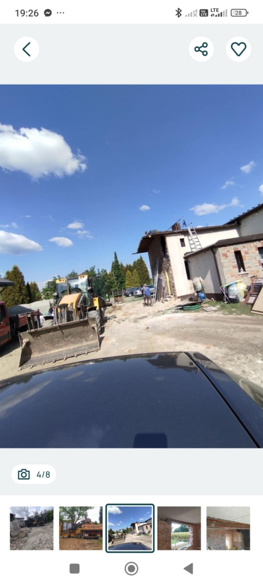 Żółta koparka na placu budowy przed częściowo rozebranym budynkiem. Widoczne gruzy, drabina oparta o ścianę i błękitne niebo z chmurami. Prace rozbiórkowe w toku.