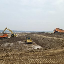 MTS MARCIN WALACHOWSKI - Żółty buldożer i koparka pracują na placu budowy, obok stoją pomarańczowe wywrotki. W tle widać drzewa i pochmurne niebo. Prace ziemne w toku.