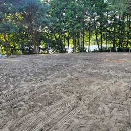 MTS MARCIN WALACHOWSKI - Przygotowany teren pod budowę z widocznymi śladami opon, wbitymi słupkami i usypanymi pryzmami piasku. W tle drzewa i jezioro. Widok na działkę przed rozpoczęciem prac.