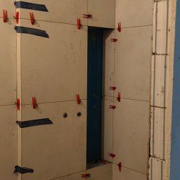 Wykończenia wnętrz Krystian Troska - Ściana w trakcie układania płytek, widoczne krzyżyki dystansowe i taśma maskująca. Fragment otworu na instalacje. Na podłodze narzędzia i resztki materiałów.