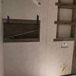 Wykończenia wnętrz Krystian Troska - Wykończona ściana w łazience z wnękami, widoczne instalacje wodne i elektryczne. Ściana pokryta beżowym tynkiem dekoracyjnym. Surowy stan podłogi.