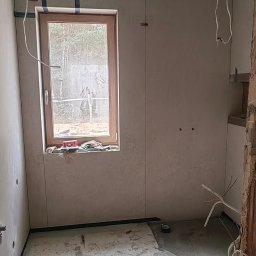 Wykończenia wnętrz Krystian Troska - Wnętrze w trakcie prac: ściany z płyt, okno z widokiem na las, widoczne przewody elektryczne. Podłoga z plamami, narzędzia na parapecie. Etap przygotowania do wykończenia.