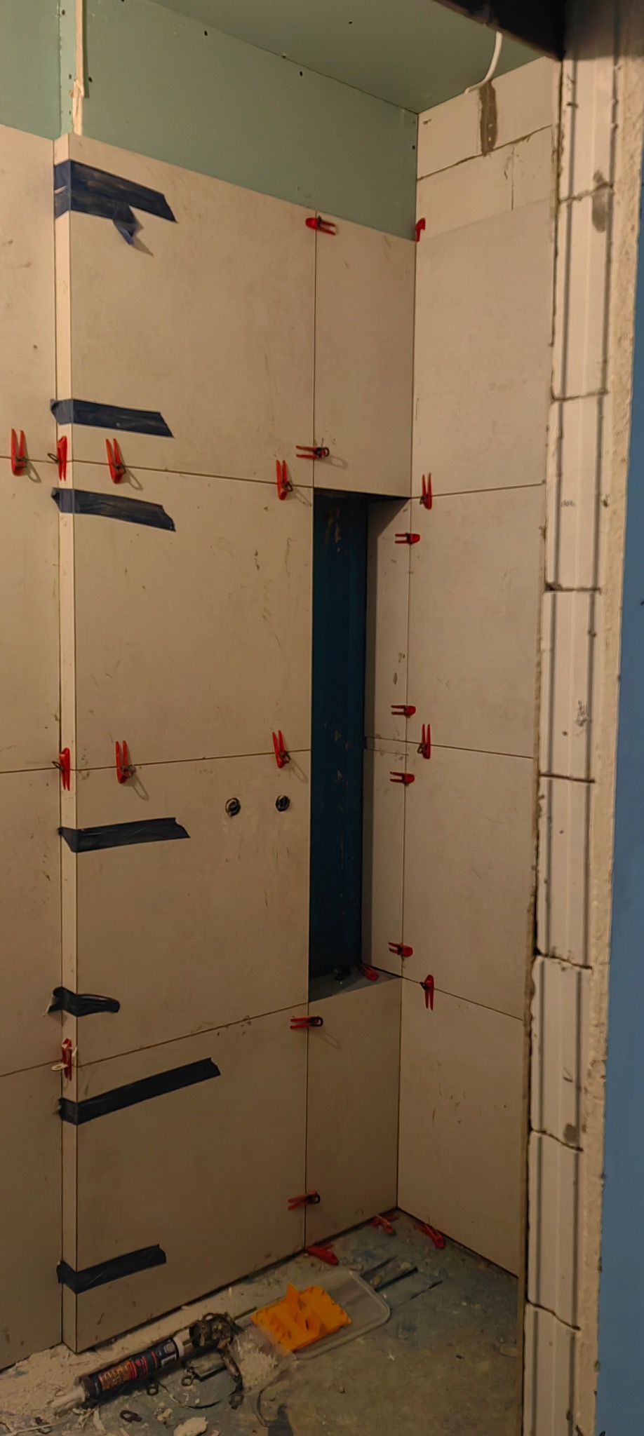 Ściana w trakcie układania płytek, widoczne krzyżyki dystansowe i taśma maskująca. Fragment otworu na instalacje. Na podłodze narzędzia i resztki materiałów.