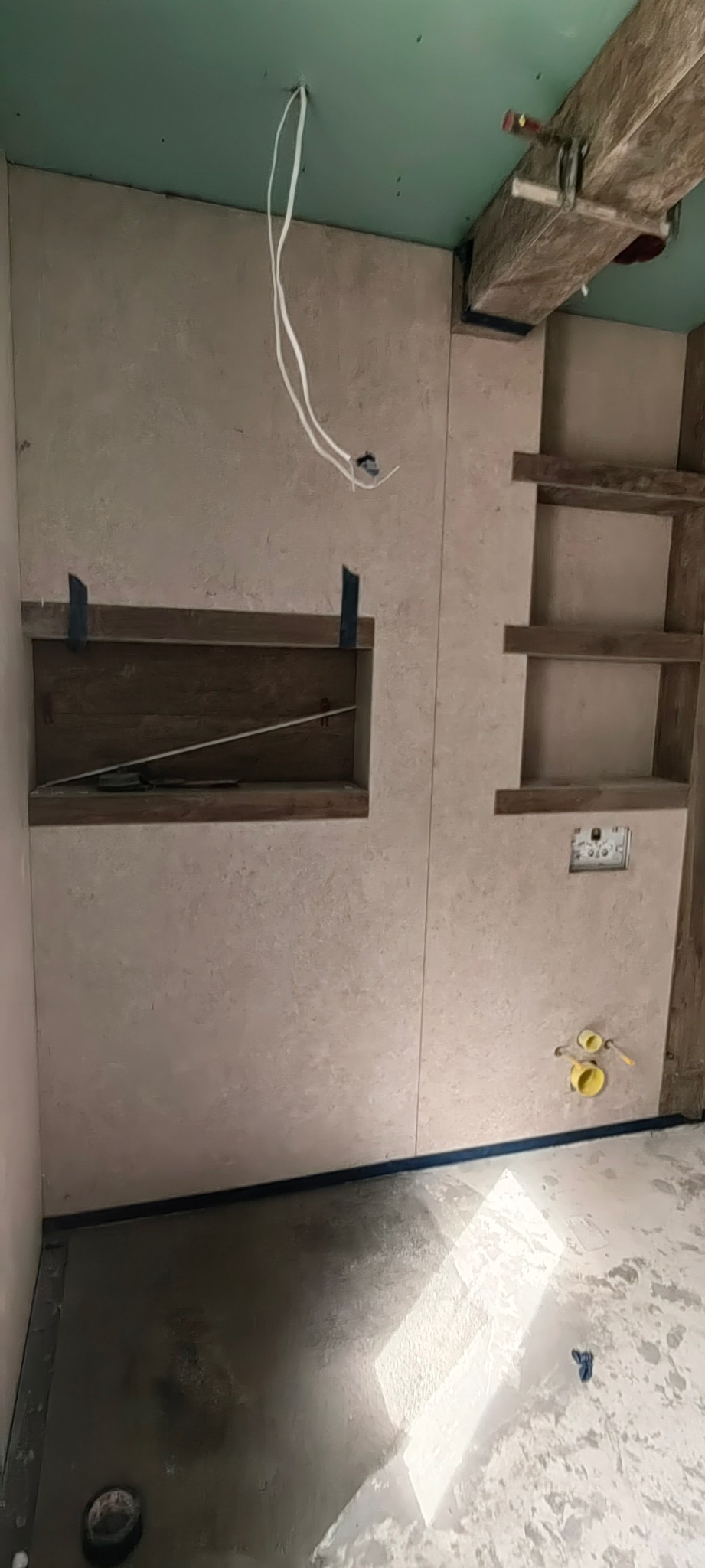 Wykończona ściana w łazience z wnękami, widoczne instalacje wodne i elektryczne. Ściana pokryta beżowym tynkiem dekoracyjnym. Surowy stan podłogi.