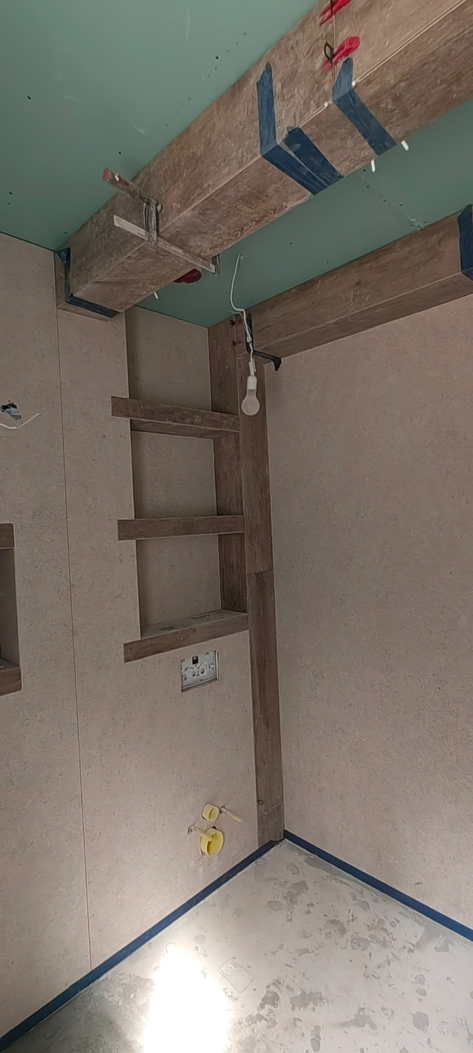 Wnętrze łazienki w trakcie prac wykończeniowych: ułożone płytki, drewniane elementy dekoracyjne, widoczne instalacje. Sufit podwieszany z płyt gipsowych.