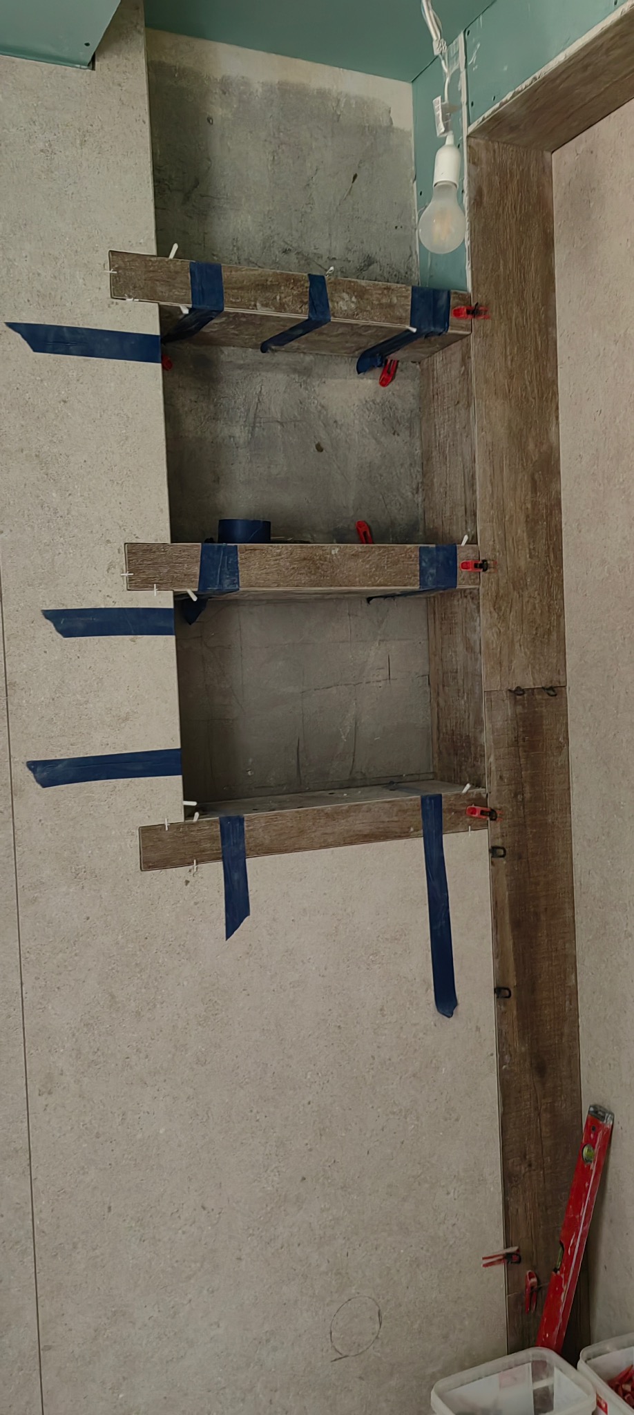 Półki wnękowe w trakcie układania płytek imitujących drewno, zabezpieczone niebieską taśmą i poziomowane czerwonymi klipsami. Widoczna surowa ściana i wisząca żarówka.