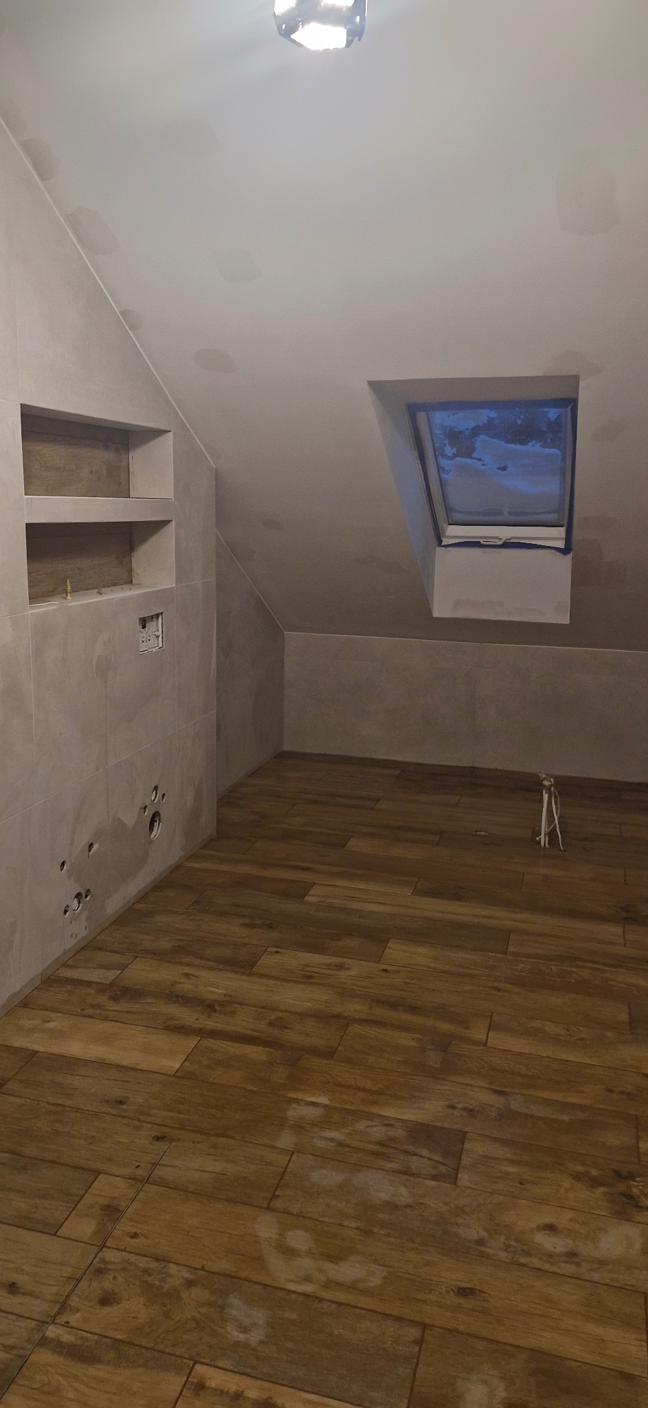 Ukończona łazienka na poddaszu z drewnopodobną podłogą, szarą glazurą i oknem dachowym. Widoczne przygotowane punkty instalacyjne na ścianie.